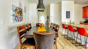 Un coin repas avec une table en bois, des chaises et un luminaire suspendu. On aperçoit en arrière-plan une cuisine avec un meuble rouge et un panneau noir.