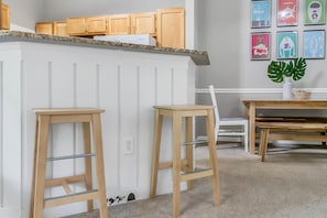 Une cuisine avec des meubles en bois, un plan de travail en granit et un îlot central blanc. On y trouve deux tabourets en bois et une table sur laquelle est posée une plante.