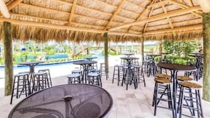 Un espace extérieur abrité, doté de tables et de chaises, avec une piscine en arrière-plan et une structure surmontée d’un toit de chaume.