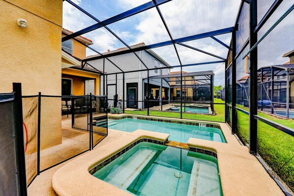 Zona de piscina con cerramiento de vidrio, espacio de descanso cubierto y vista de edificios cercanos.