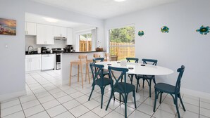 A modern kitchen with white cabinets, a dining area with a round table and teal chairs, and a large window with blinds. - Pearl Villa (Kissimmee)