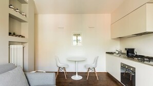 Eine kompakte Küche mit weißen Schränken, einem Herd und einer Mikrowelle. Über der Spüle befindet sich ein kleines Fenster und in der Mitte steht ein weißer Tisch mit zwei Stühlen.
