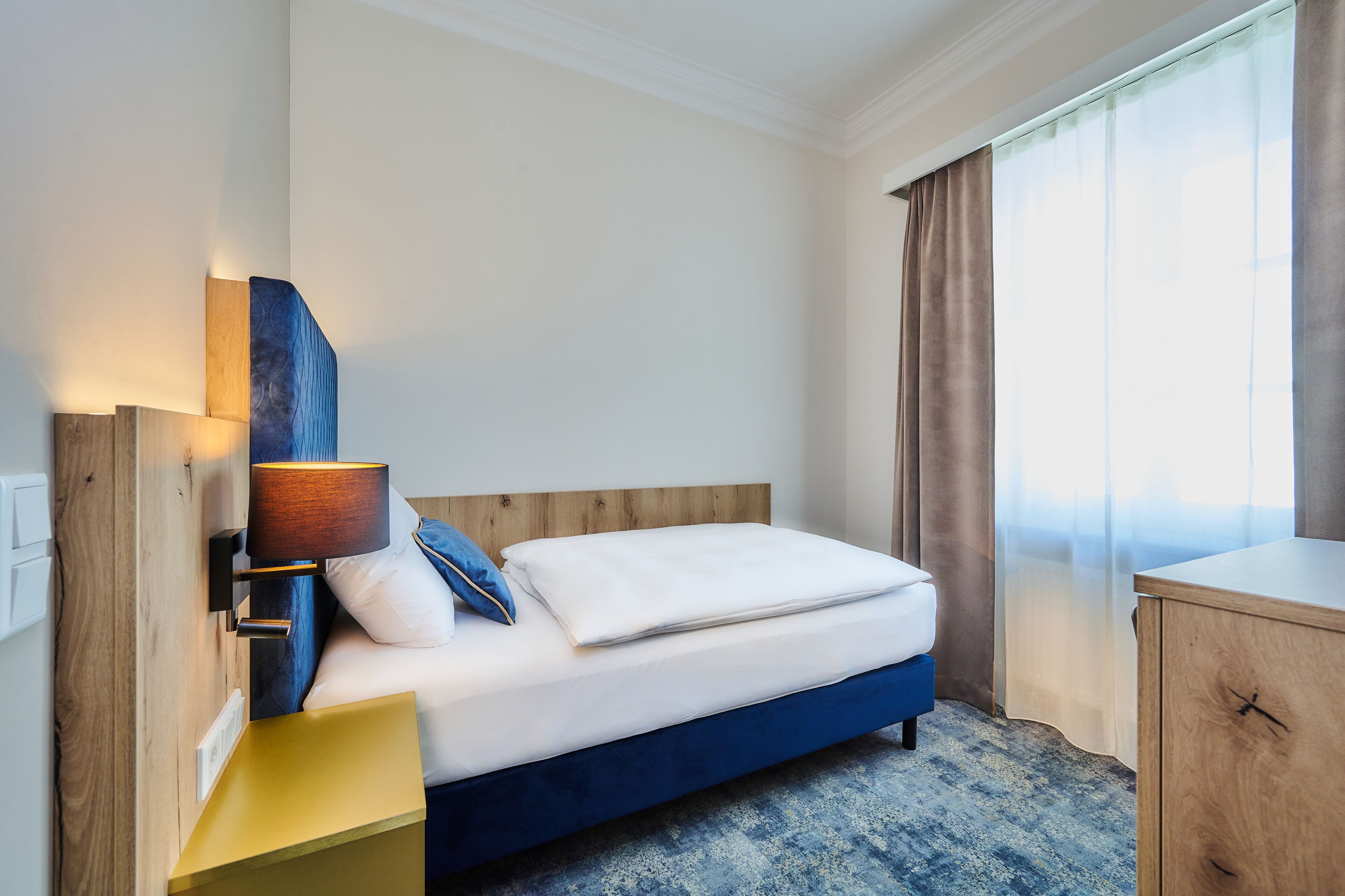 Quarto de hotel com cama, mesa de cabeceira, abajur, cabeceira de madeira, janela com cortinas e um armário de madeira.