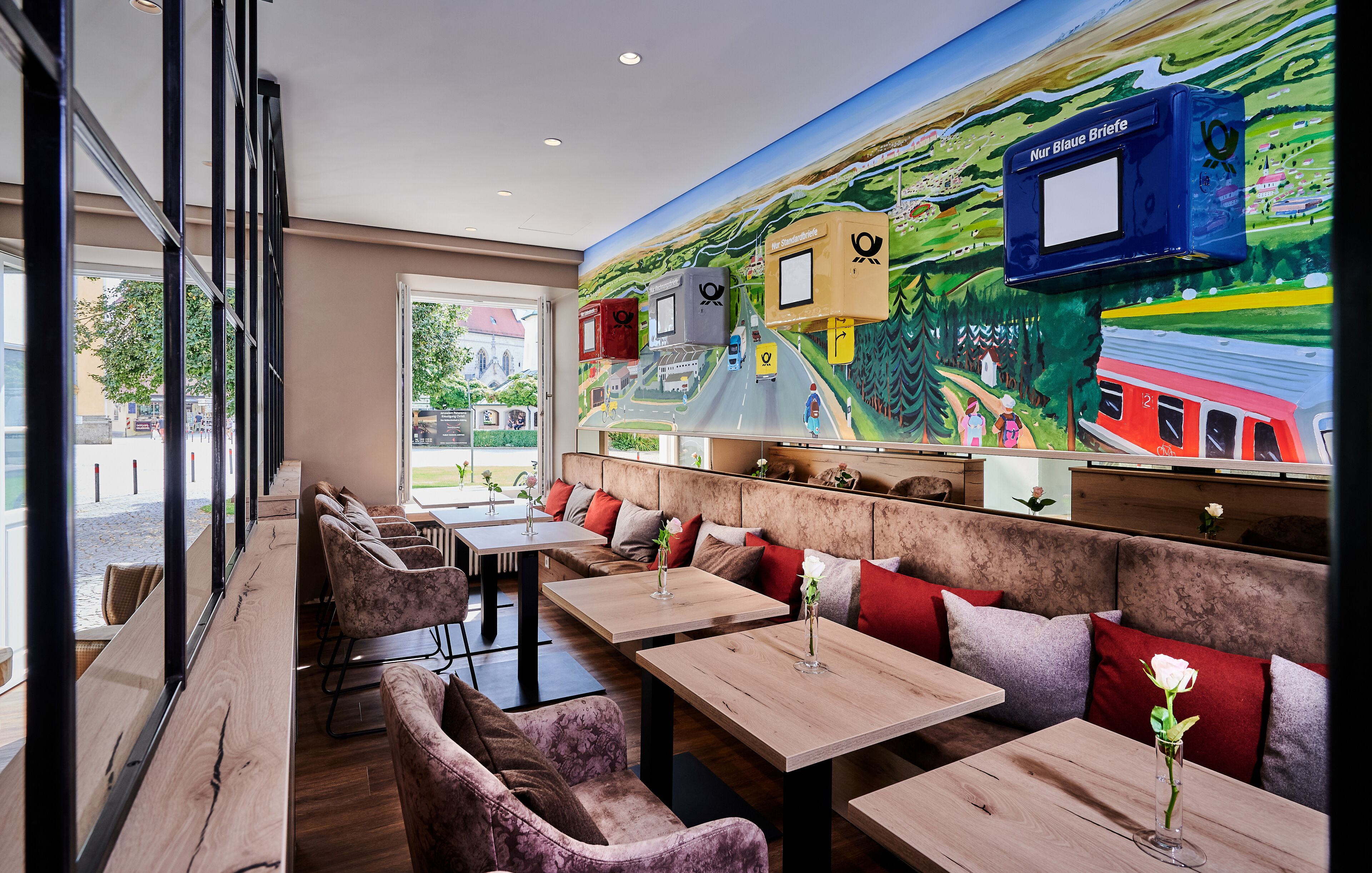 A restaurant with wooden tables and cushioned booths, a mural of a train on the wall, and a view of the street outside.
