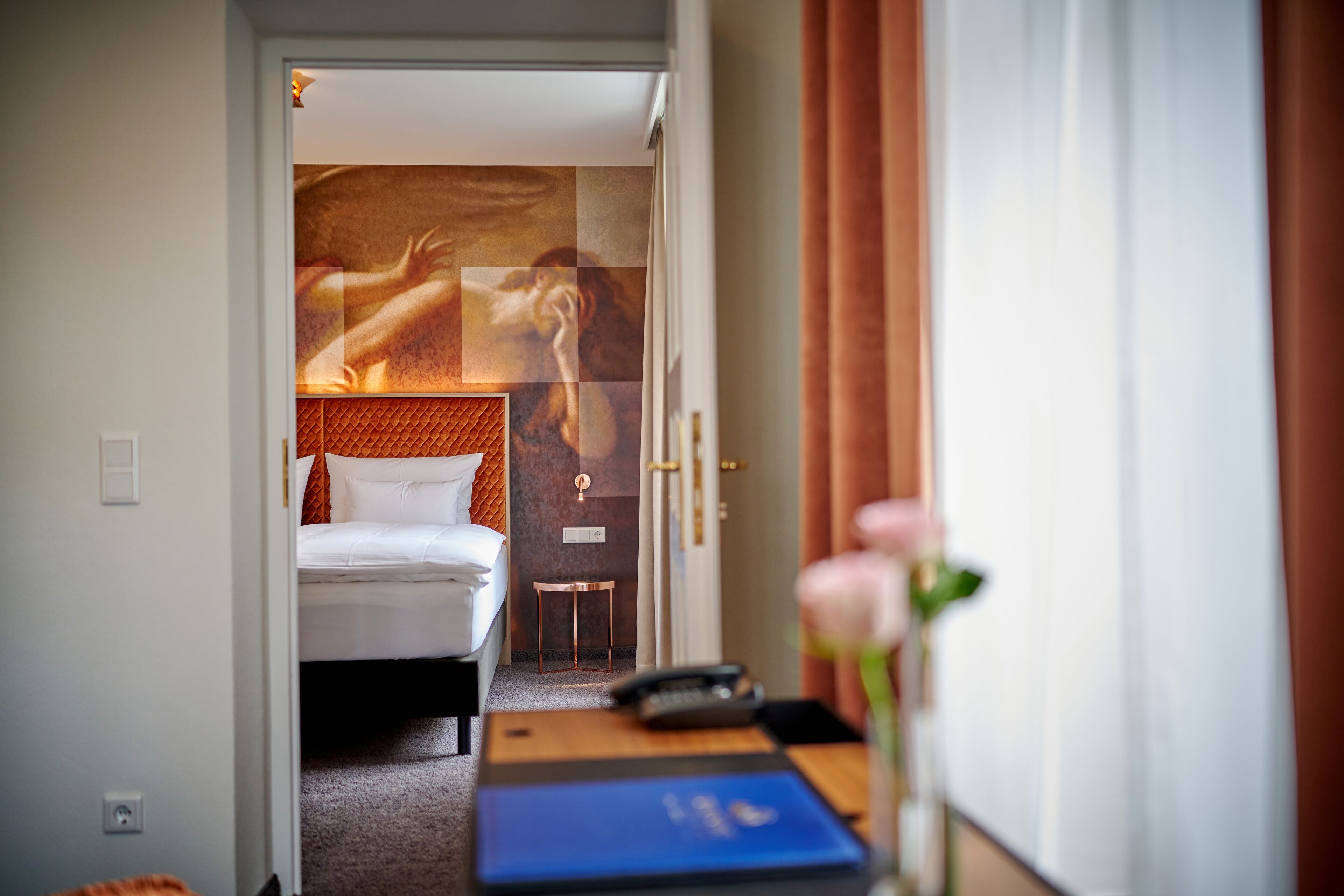 Et hotelværelse med en seng, et lille bord med en telefon og en bog, samt en væg med et stort kunstværk.