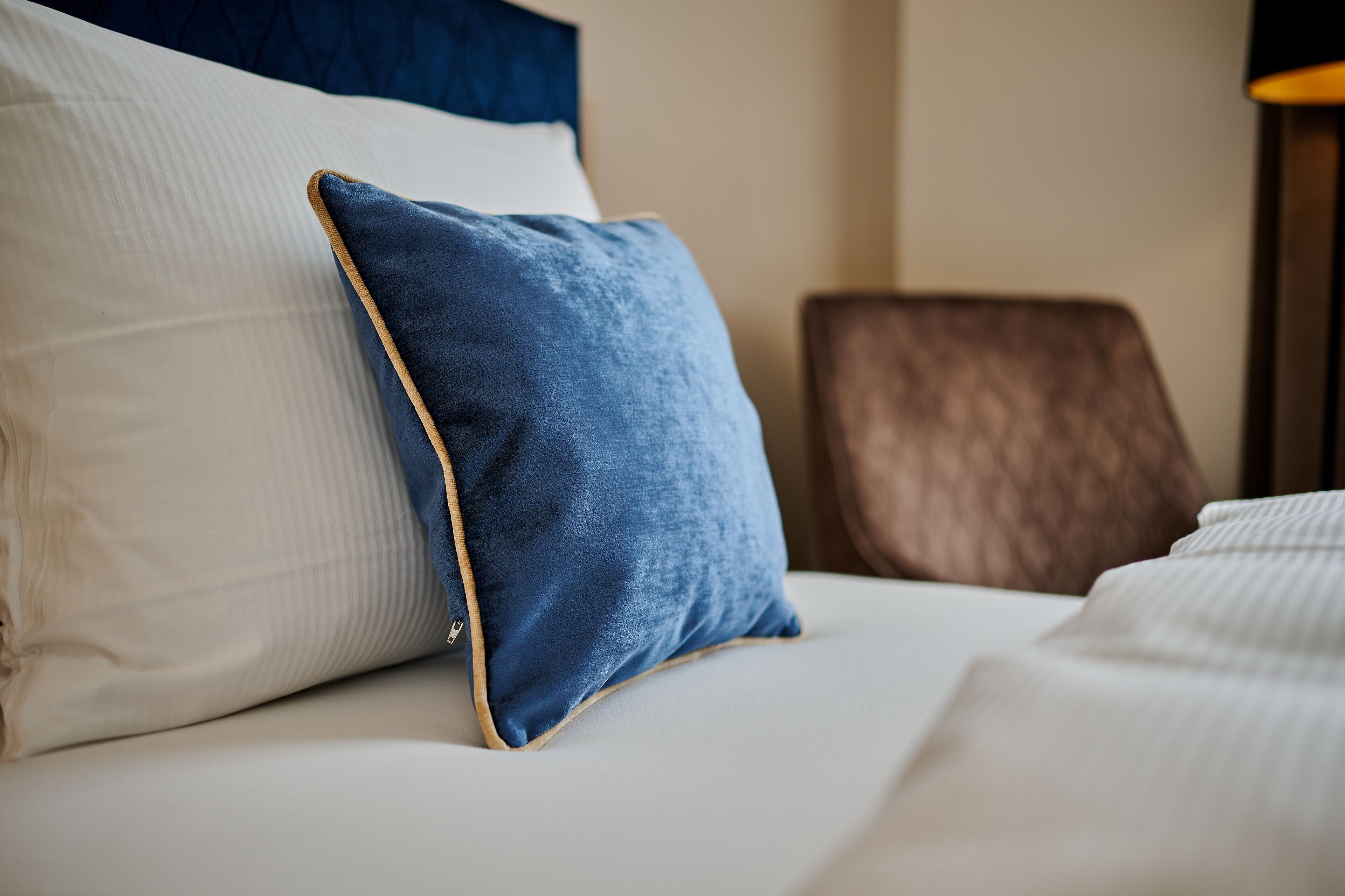 A bed with a white and a blue pillow, a brown chair, and a lamp.
