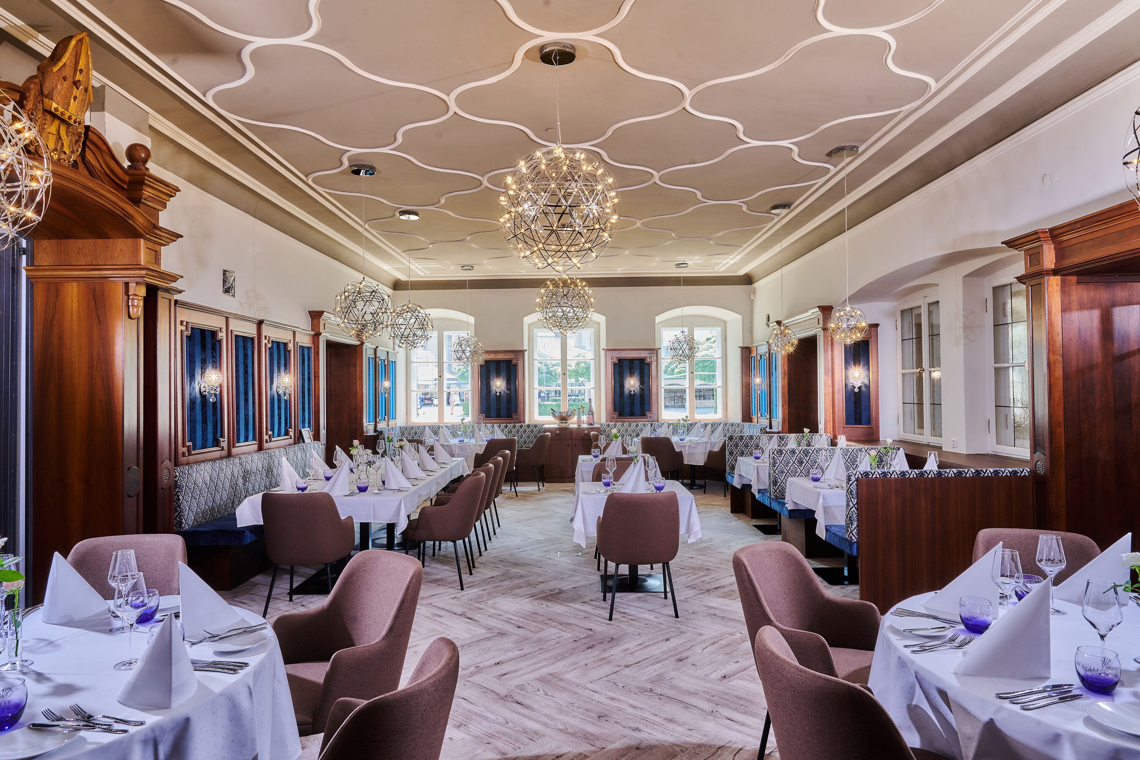 Ein Restaurant mit hohen Decken, aufwendigen Kronleuchtern und klassischem Interieur. Die Tische sind mit weißen Tischdecken und violettem Glas bestellt.