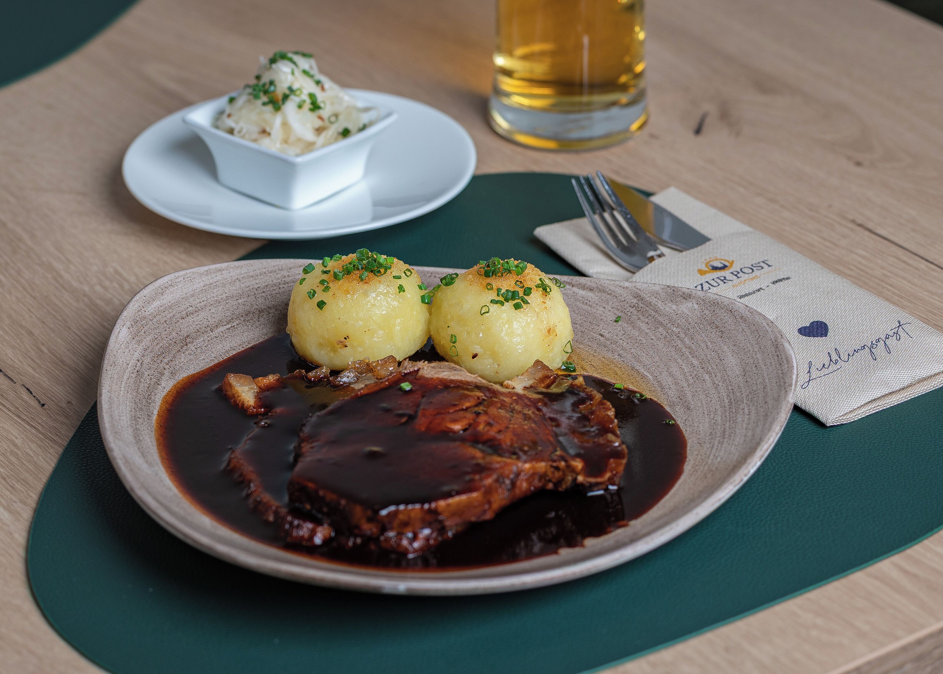 A plate of food with a meat dish, two round potatoes, and a small bowl of sauce on a table.