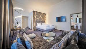 A modern hotel room with a large bed, a sofa, a coffee table, a TV, and a bathroom with a large mirror and sink. - Hotel Zur Post Altötting (Altötting)