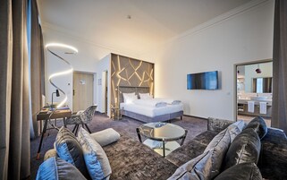 A modern hotel room with a large bed, a sofa, a coffee table, a TV, and a bathroom with a large mirror and sink.