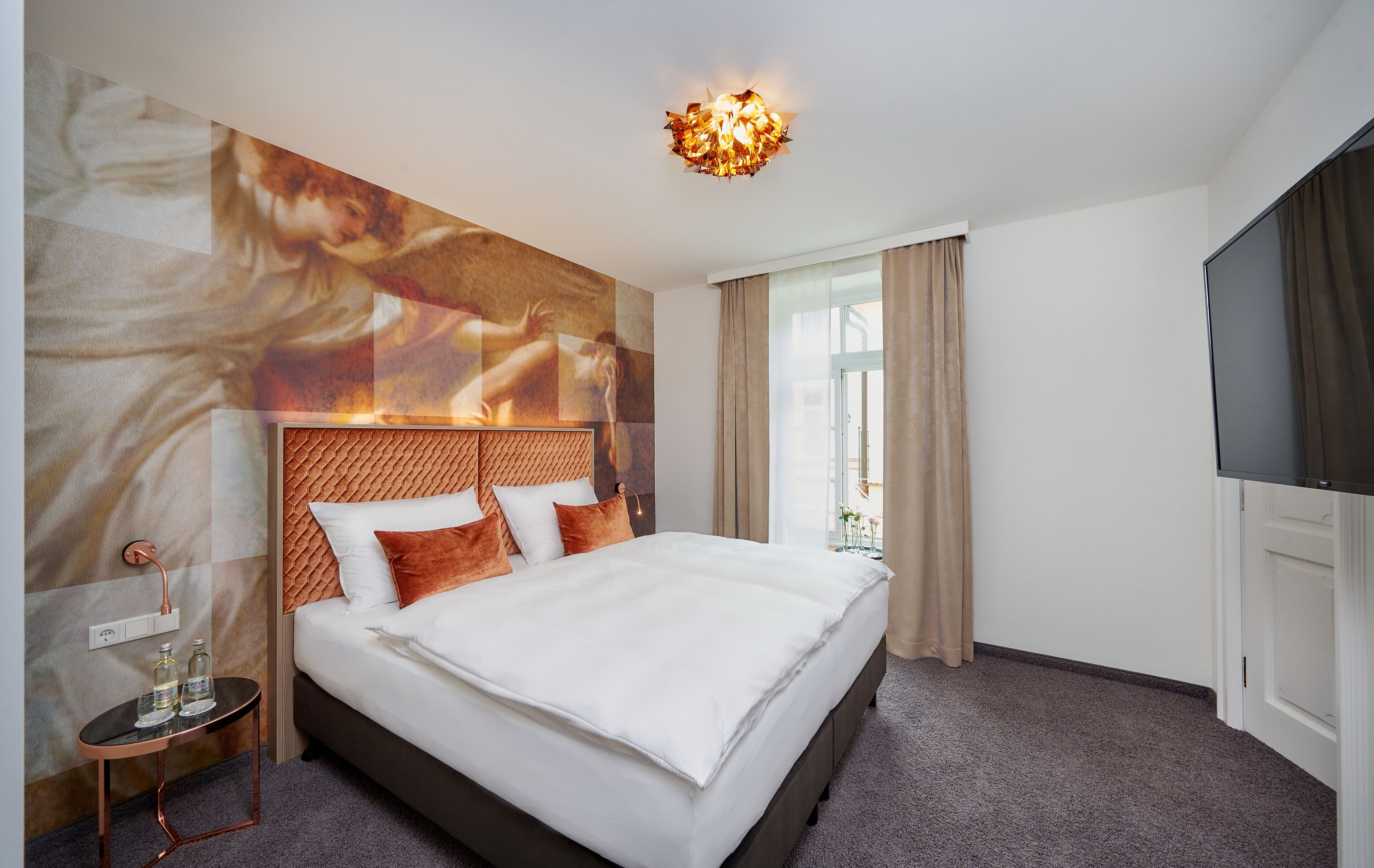 Une chambre d’hôtel avec un grand lit, une tête de lit au motif texturé, un téléviseur fixé au mur et une fenêtre avec des rideaux.