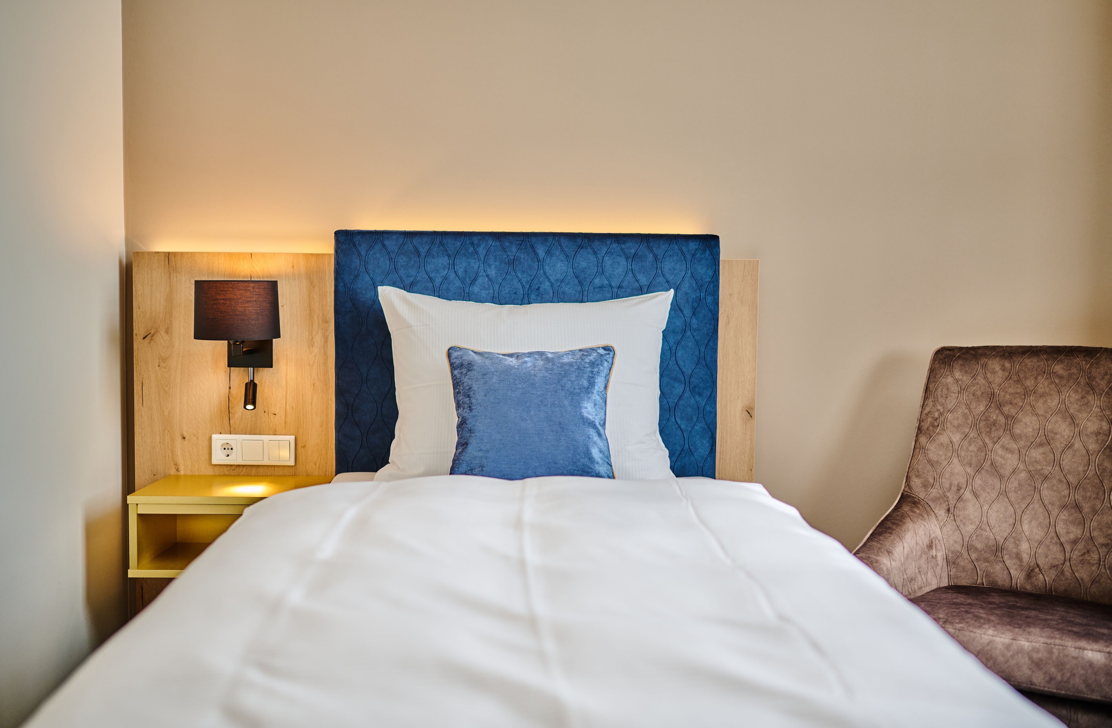 A neatly made bed with a blue headboard, a bedside lamp, and a brown armchair.