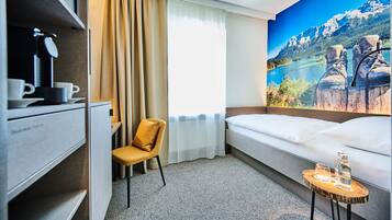 Ein Hotelzimmer mit Bett, Schreibtisch mit Kaffeemaschine, Stuhl, kleinem Tisch mit Gläsern und Flaschen, Fenster mit Vorhängen und einem Wandgemälde von einer Berglandschaft.