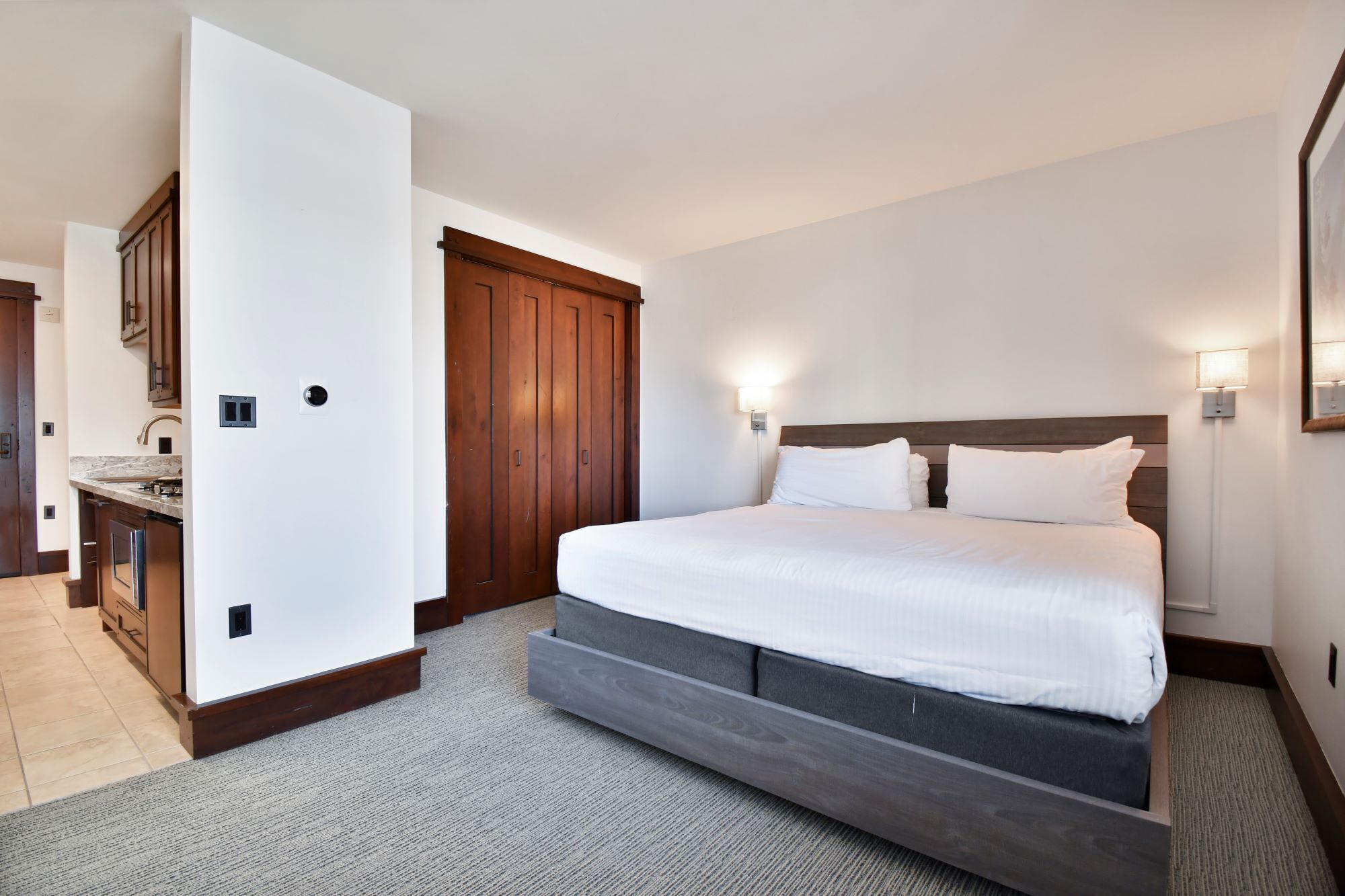 A hotel room with a bed, a wooden wardrobe, a bathroom with a shower, and a small kitchen area.