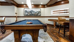 A pool table with balls, a pool cue, and a hanging light fixture in a room with wooden paneling and a framed picture on the wall. - Ski-In Ski-Out Condo at Deer Valley (Park City)