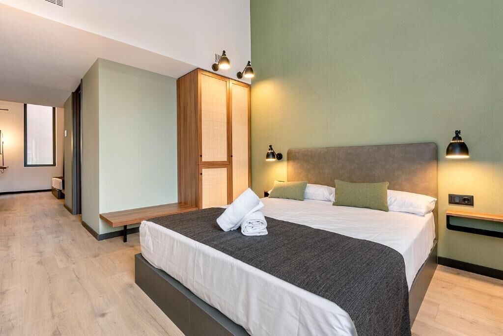 A modern hotel room with a large bed, bedside tables, a wardrobe, and a small seating area.