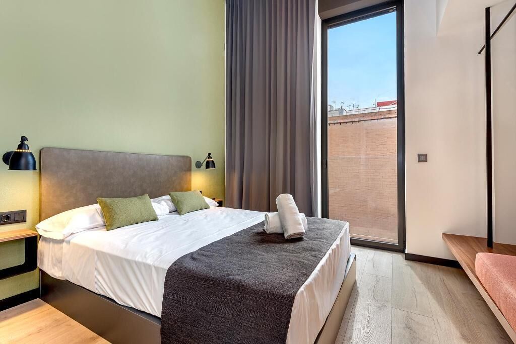 A modern hotel room with a bed, a nightstand, a lamp, and a view of the outside.