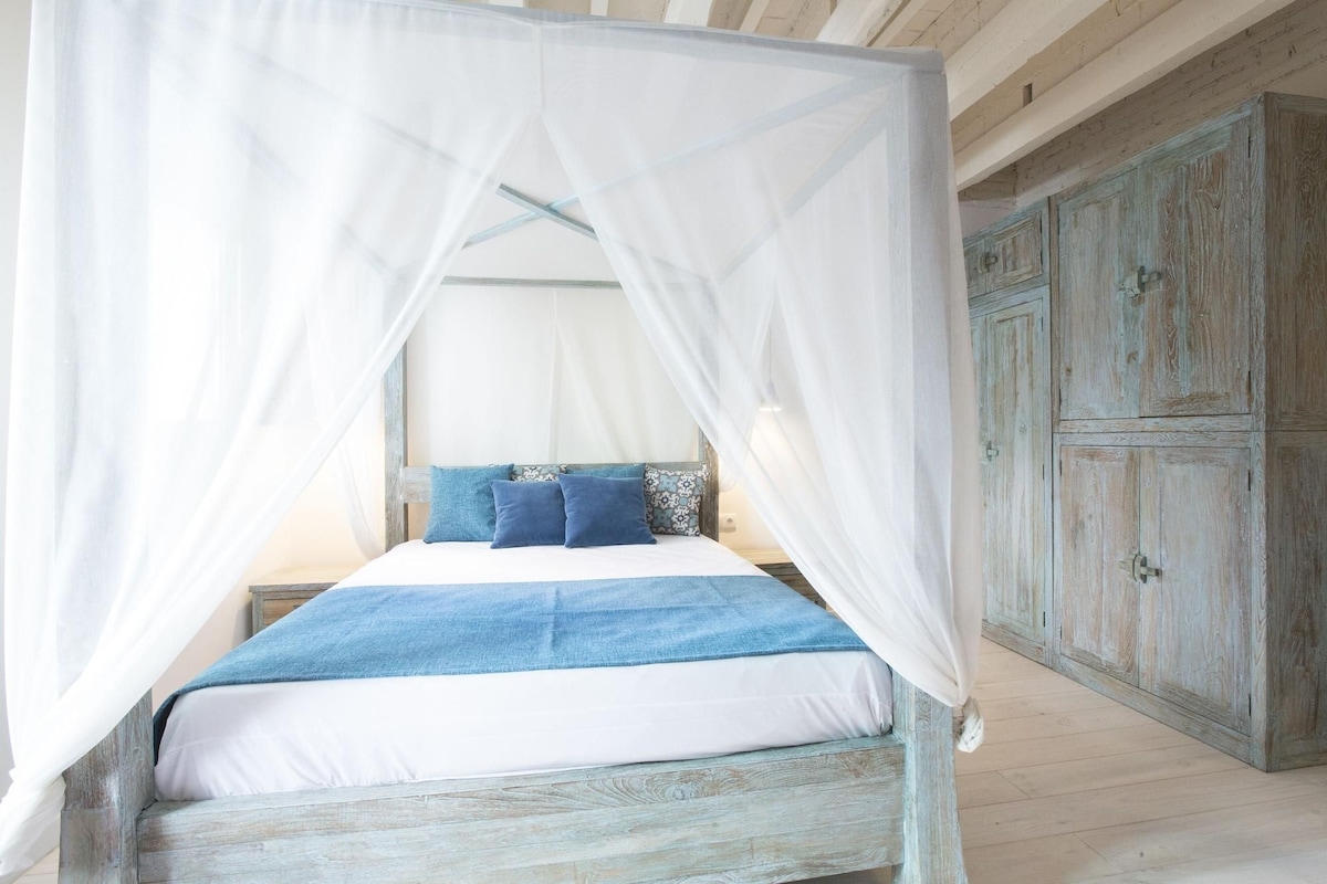 Ein Bett mit einem weißen Moschee-Vorhang, blauer Bettwäsche und einem Holzkopfende.