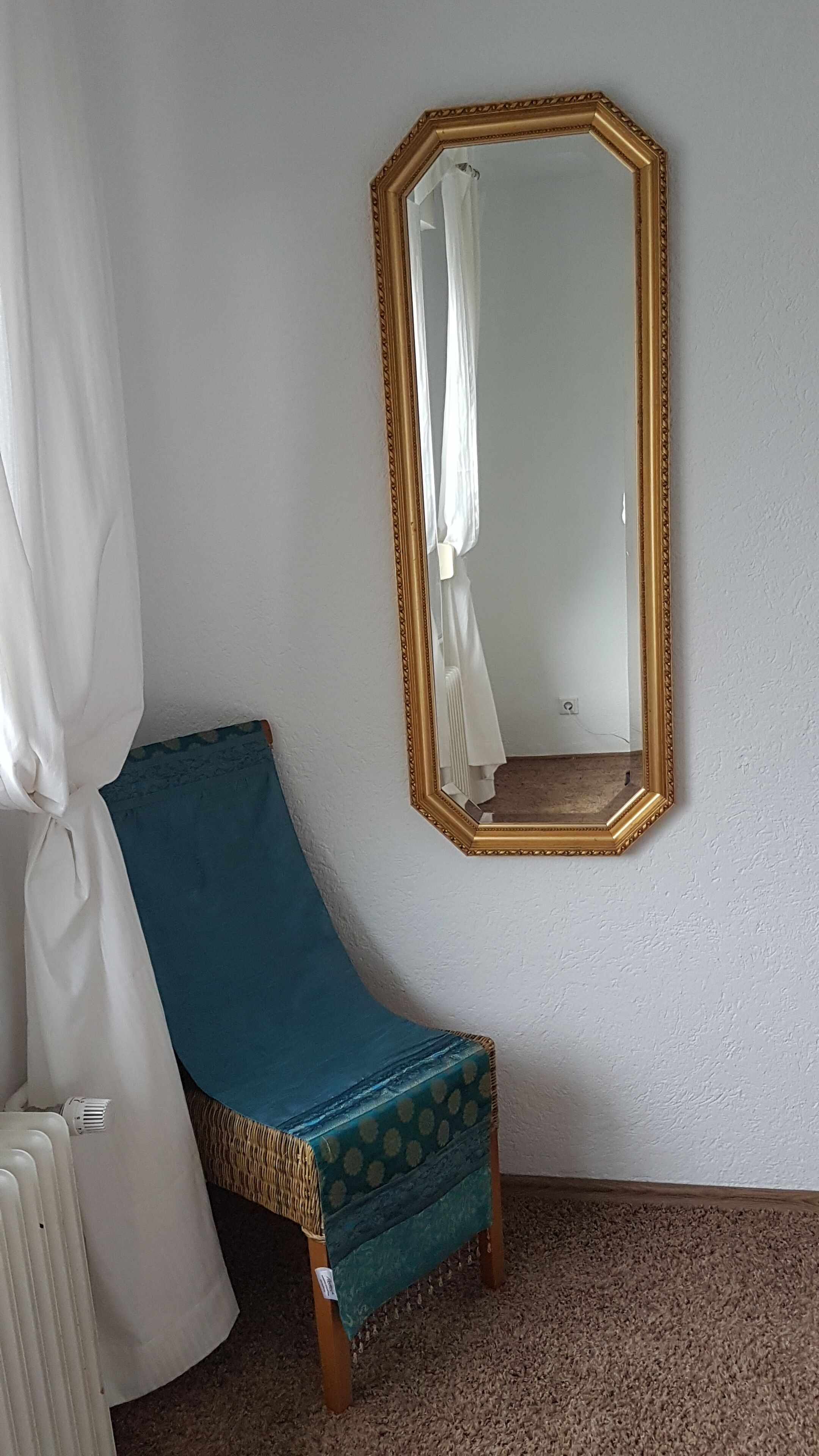 Una habitación con una cama individual, un espejo en la pared, una cortina blanca y una alfombra marrón.