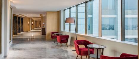 A hotel corridor with red chairs, a lamp, and large windows offering a view of the city.
