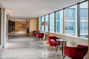 Un pasillo de hotel con sillas rojas, una lámpara y ventanas amplias que dan a vistas de la ciudad.