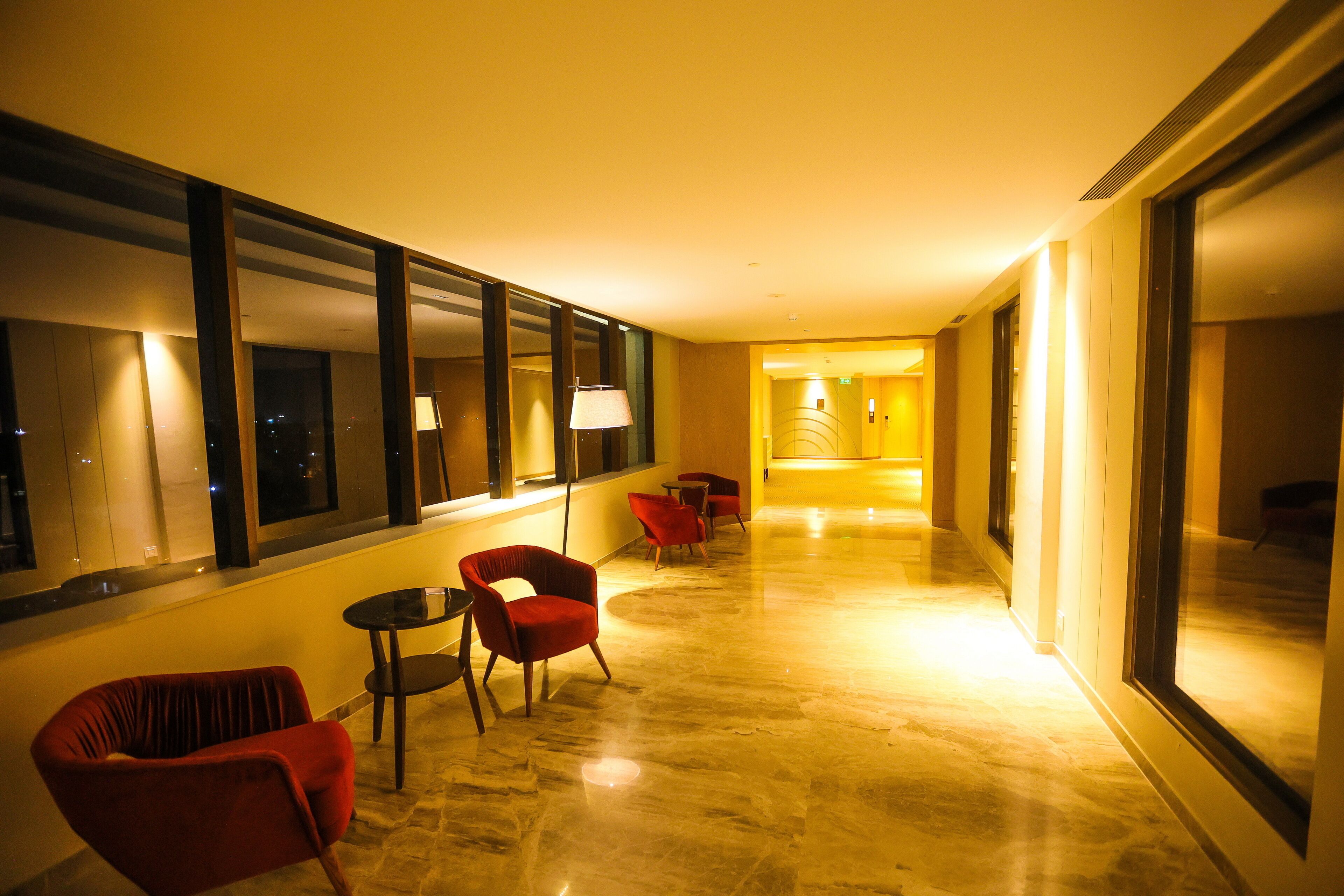A hotel corridor with a marble floor, red chairs, and a lamp near the window.