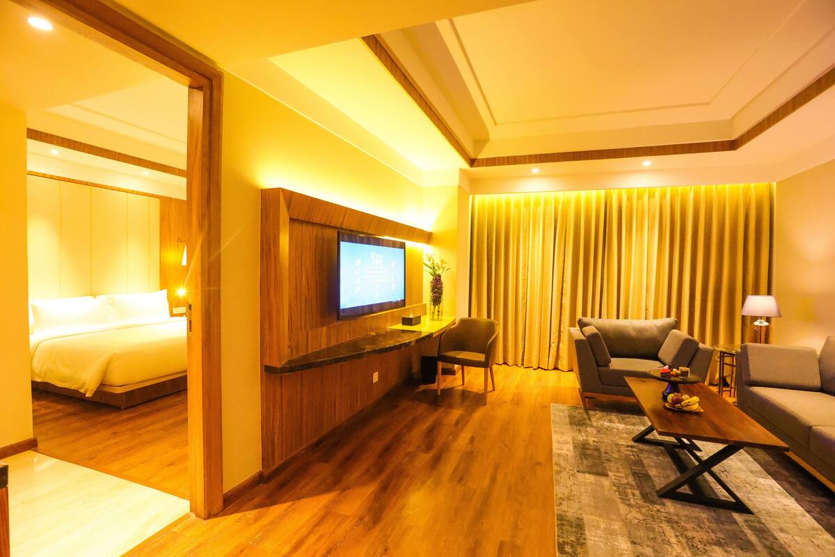 A hotel room with a wooden floor, a bed, a TV mounted on the wall, a sofa, a coffee table, and a curtain.