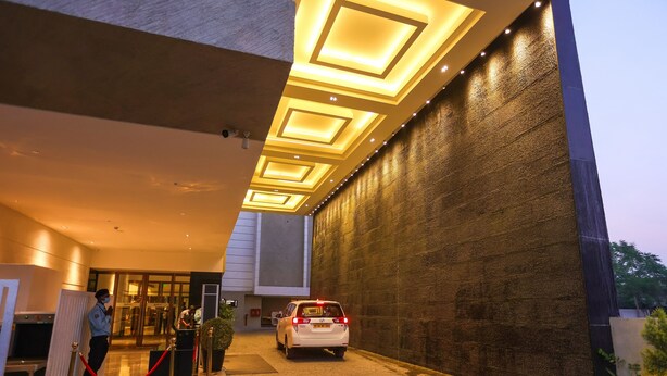 Une entrée moderne d’un immeuble, avec une voiture blanche garée devant, un agent de sécurité et une barrière en corde rouge.