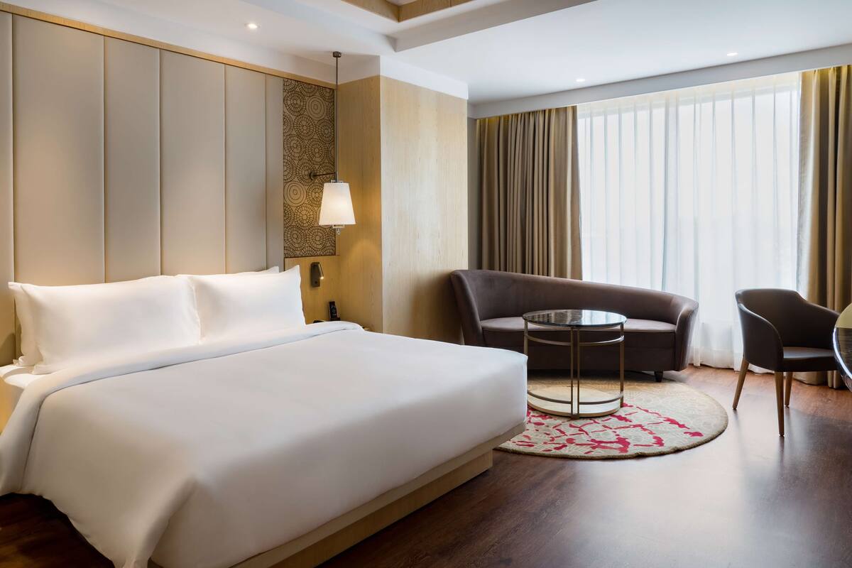 A hotel room with a large bed, a sofa, a small table, and a view of the outdoors through large windows.