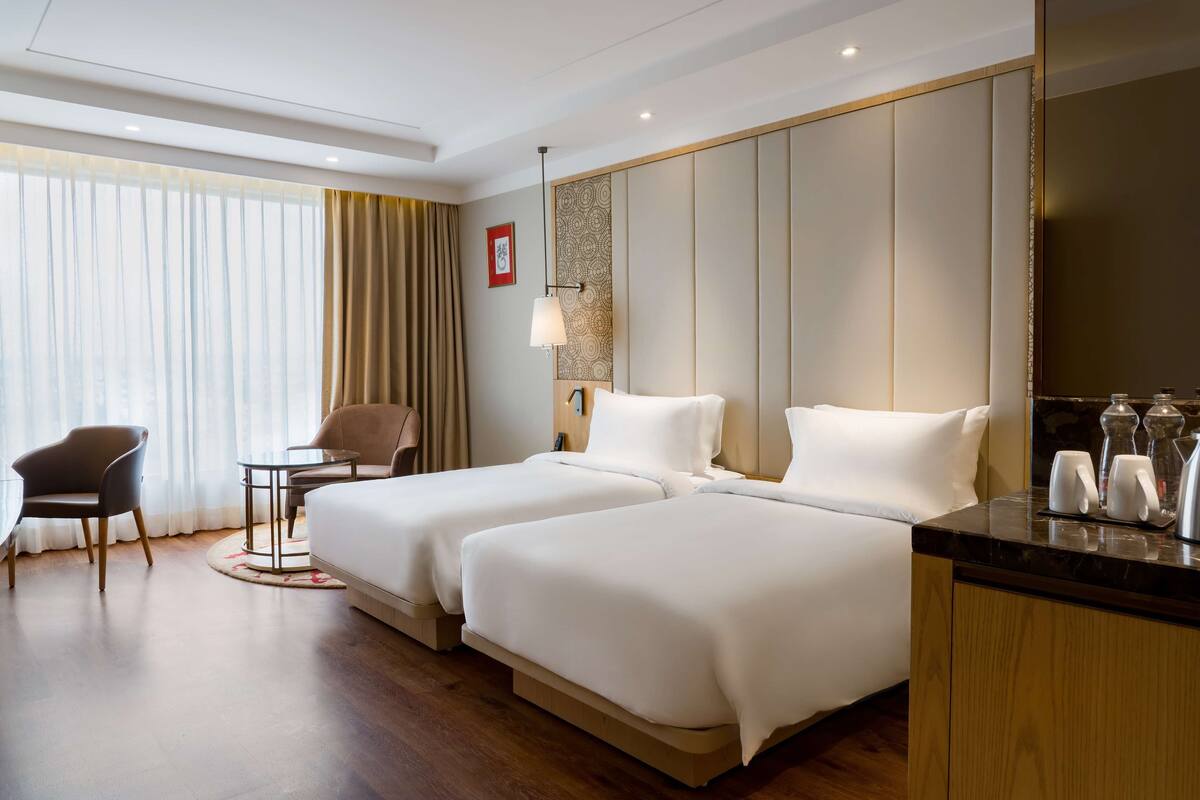 A hotel room with two beds, a desk, a chair, a lamp, and a large mirror.