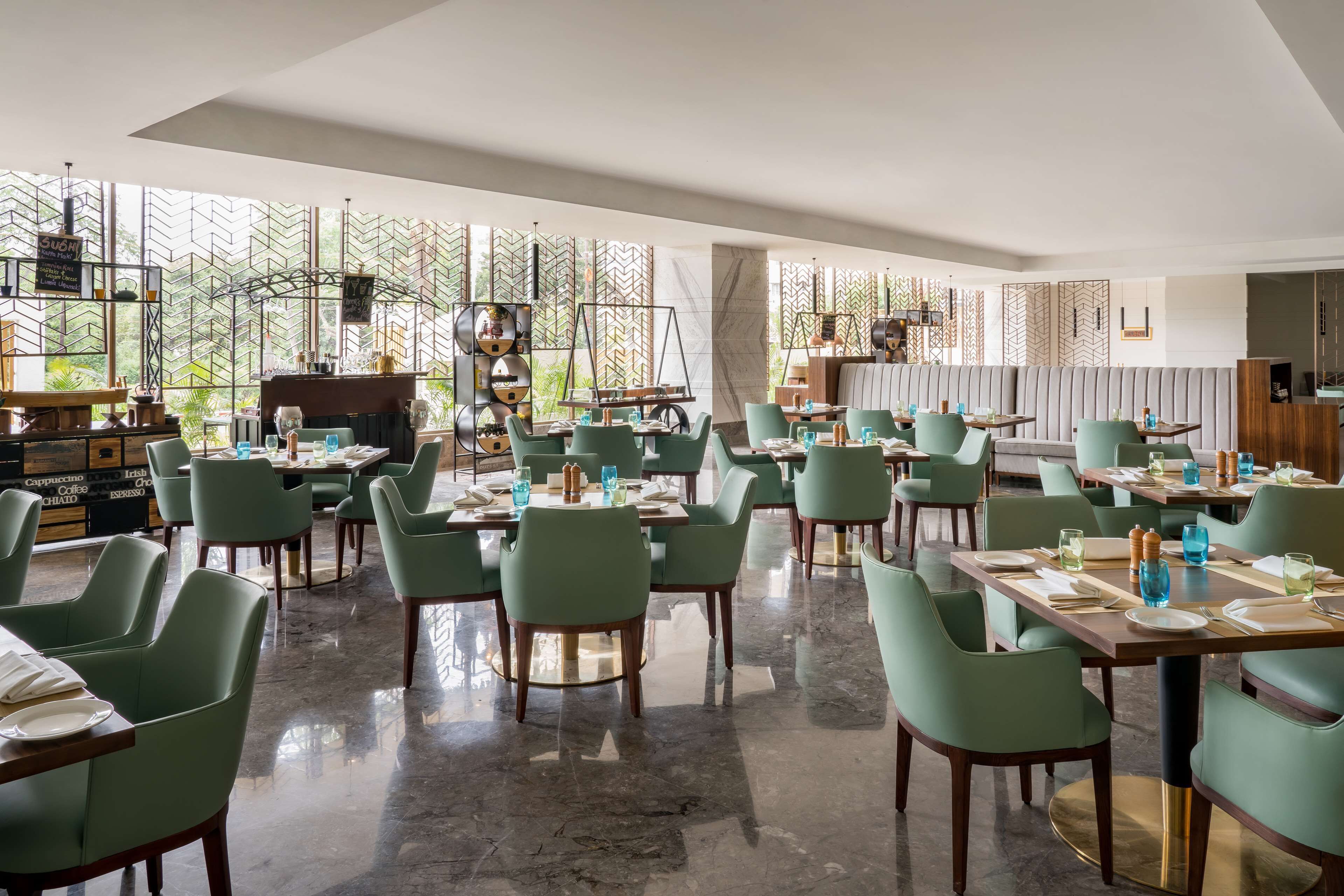 A modern restaurant with green chairs and tables, large windows, and a decorative wall.