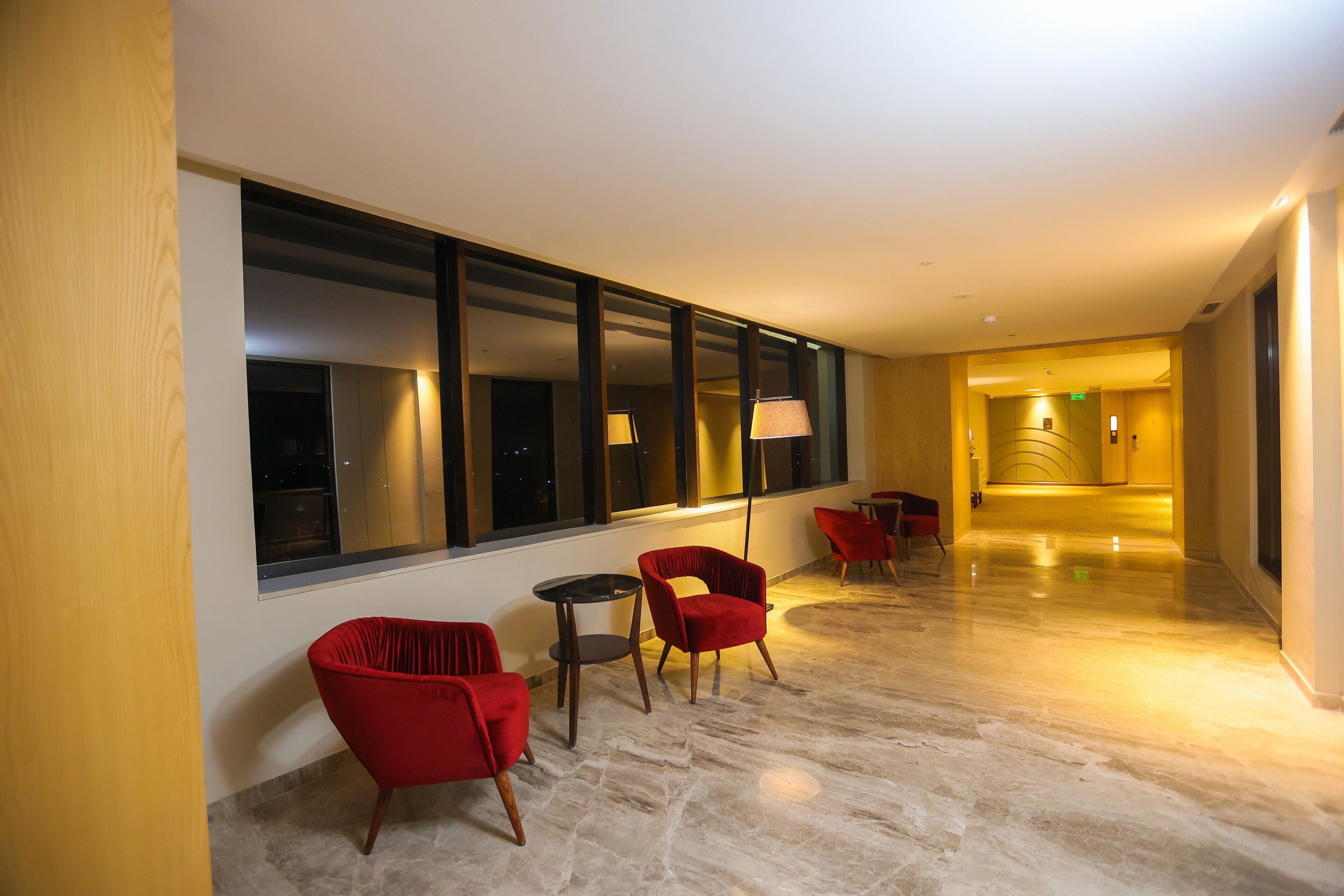 Un pasillo de hotel con sillas rojas, una mesita y ventanas amplias.