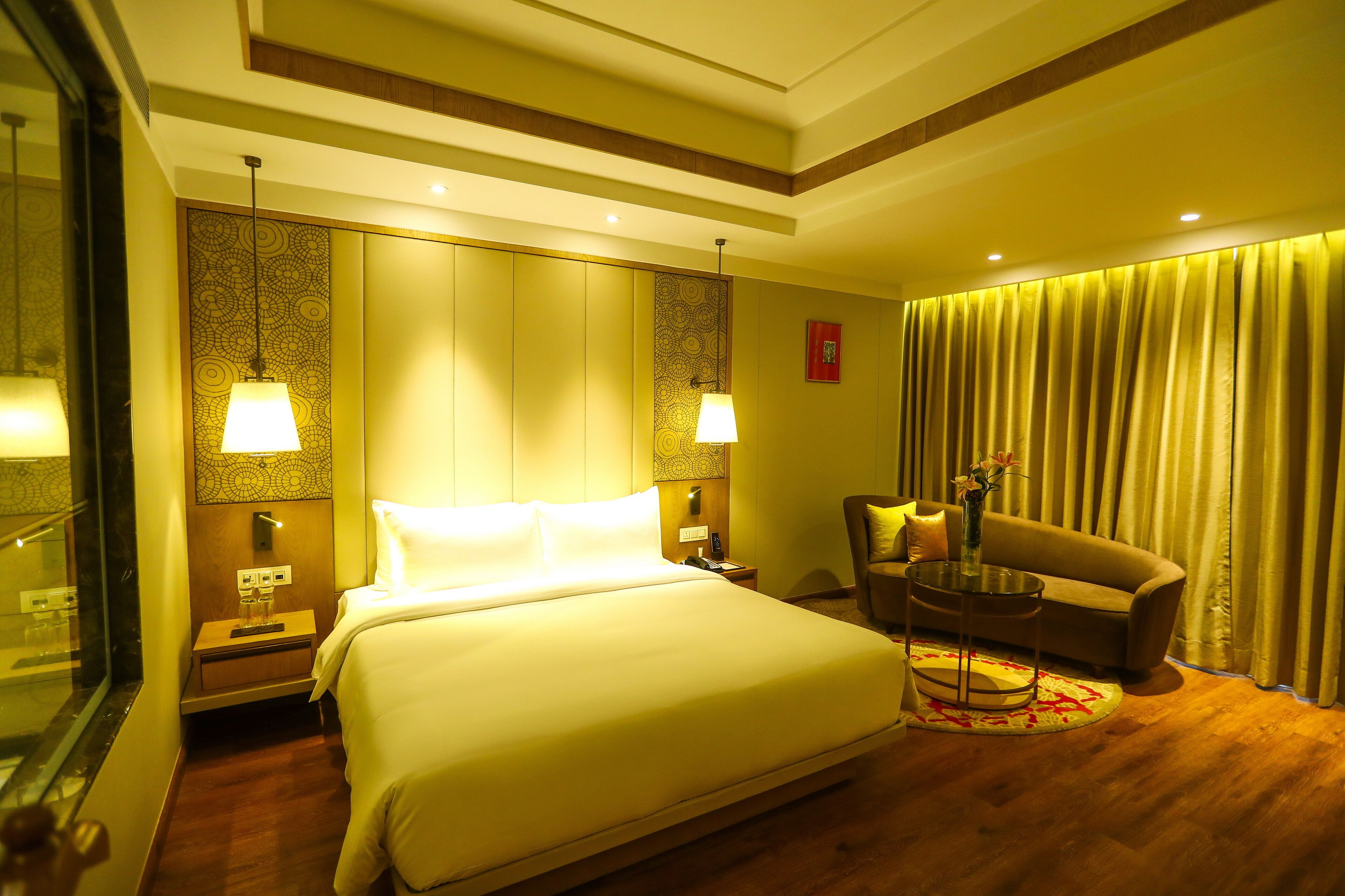 A hotel room with a large bed, a sitting area with a chair and a small table, a nightstand, and a mirror.