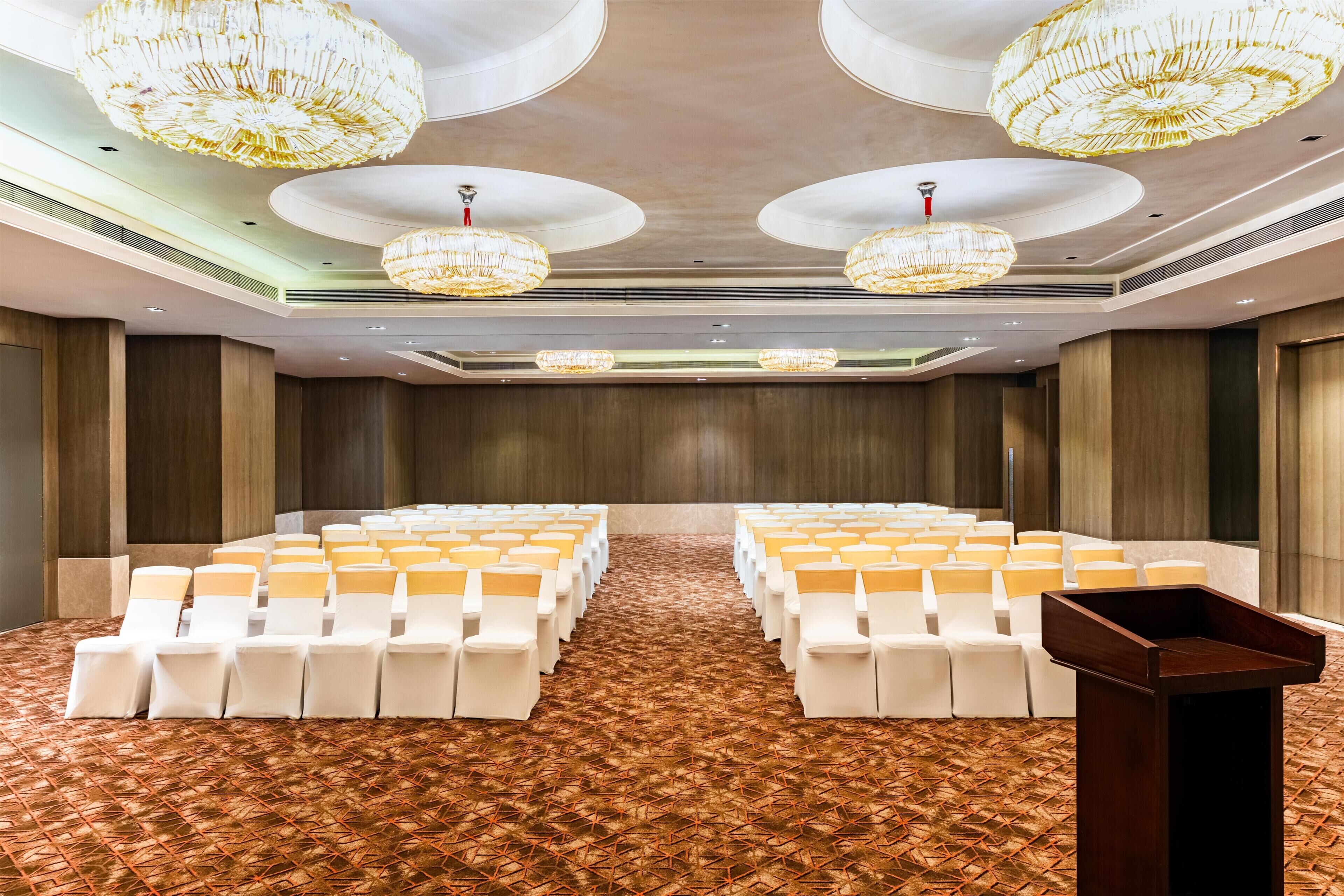 A conference room with rows of white chairs, a patterned carpet, and large chandeliers.