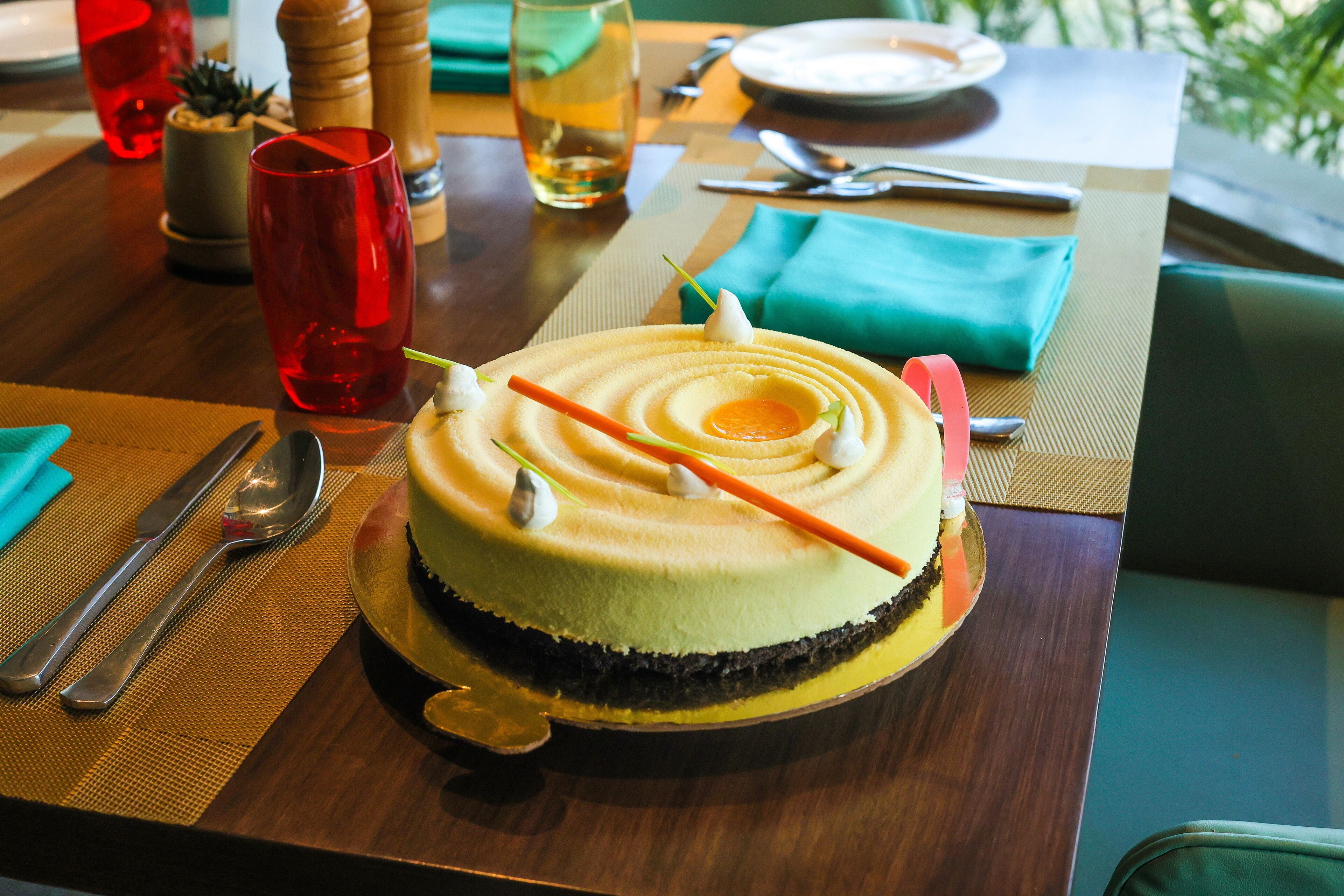 Un postre sobre una mesa de madera con un mantel individual, rodeado de cubiertos y un vaso de bebida.