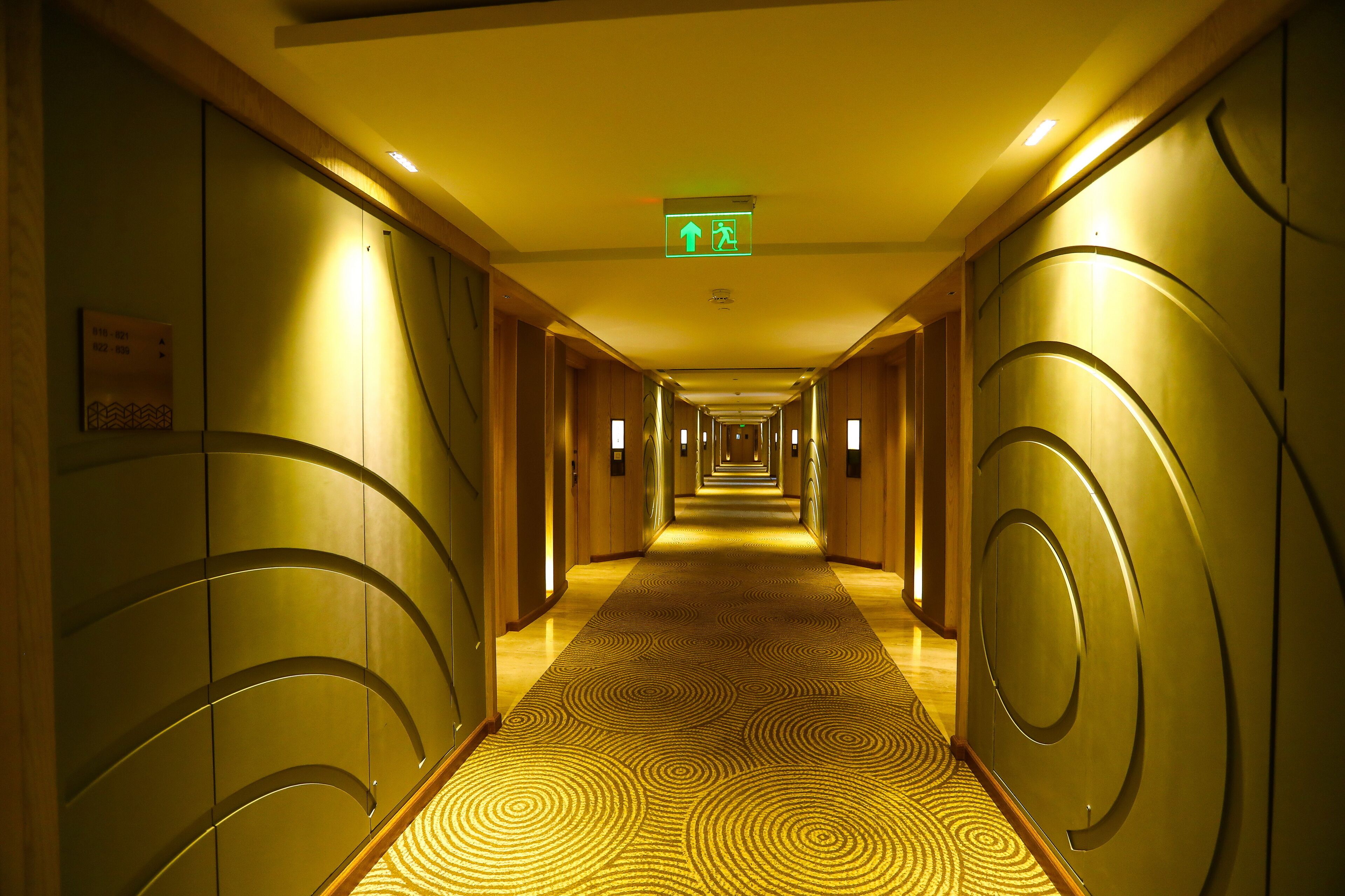 Un pasillo de hotel con alfombra estampada, paredes de madera y un letrero de salida de emergencia verde.