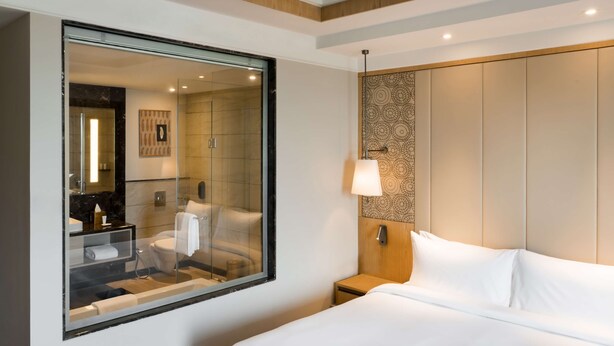 A hotel room with a large mirror, a bed with white linens, a nightstand with a lamp, and a bathroom visible through the mirror. - Radisson Bhopal (Bhopal)