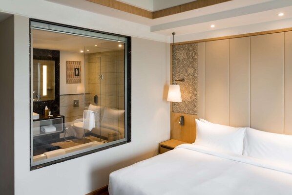 Habitación de hotel con un espejo grande, una cama con sábanas blancas, una mesita de noche con una lámpara y un baño visible a través del espejo.