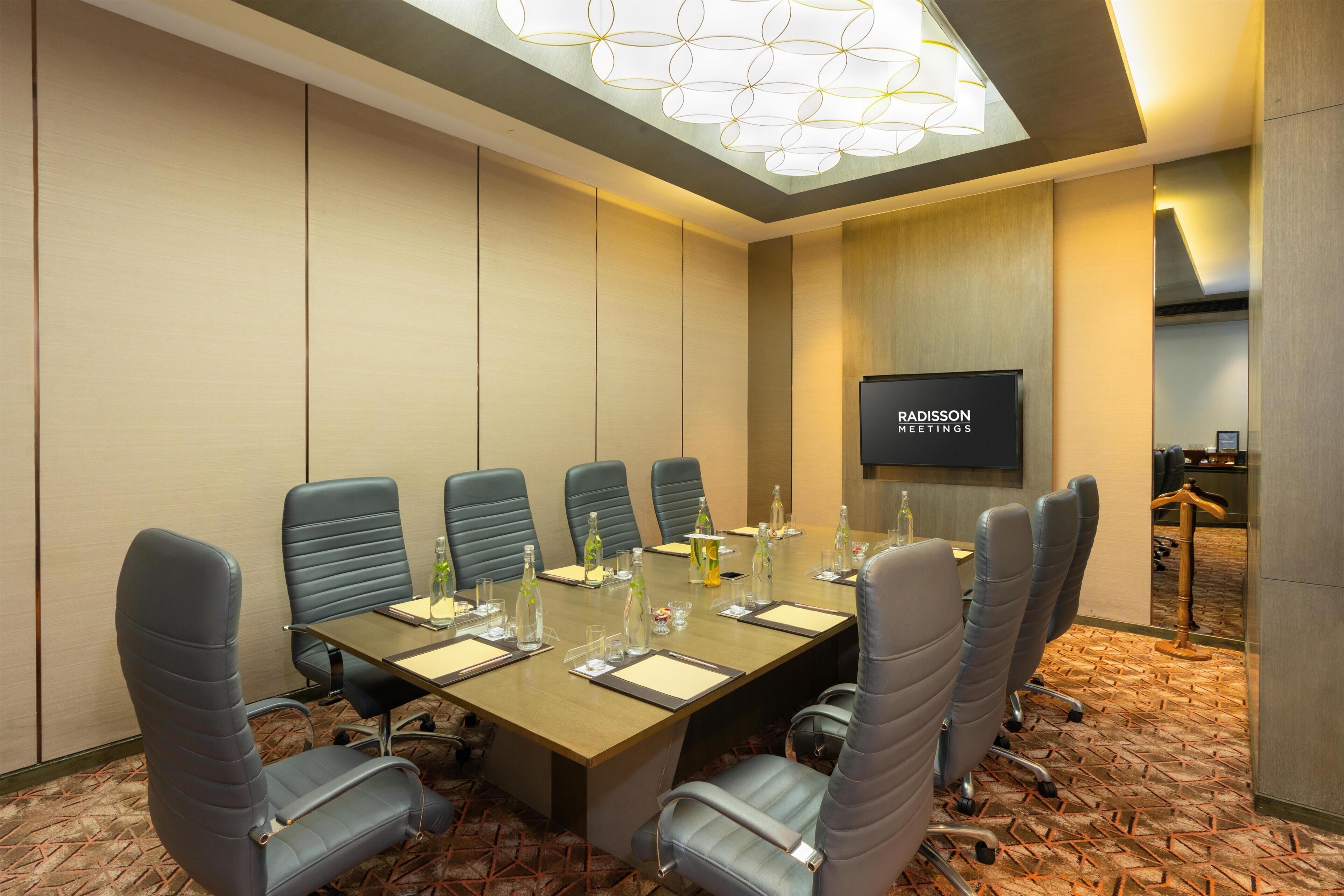 A conference room with a long table, six chairs, a television screen, and a patterned carpet.