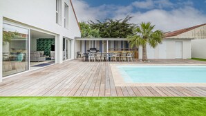 Een modern huis met een houten terras, een zwembad en een overdekte zitplek buiten.