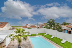 Zwembad met witte ligbedden, een palmboom en uitzicht op omliggende huizen.