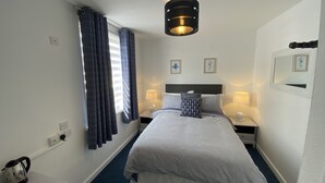 Et hotelværelse med seng, to sengeborde med lamper, en loftlampe, et vindue med gardiner, et spejl på væggen og et lille bord med elkedel og kopper.
