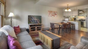 Una sala de estar moderna con un sofá gris, una mesa de centro de madera y un televisor. Hay una zona de cocina con armarios blancos y electrodomésticos de acero inoxidable.