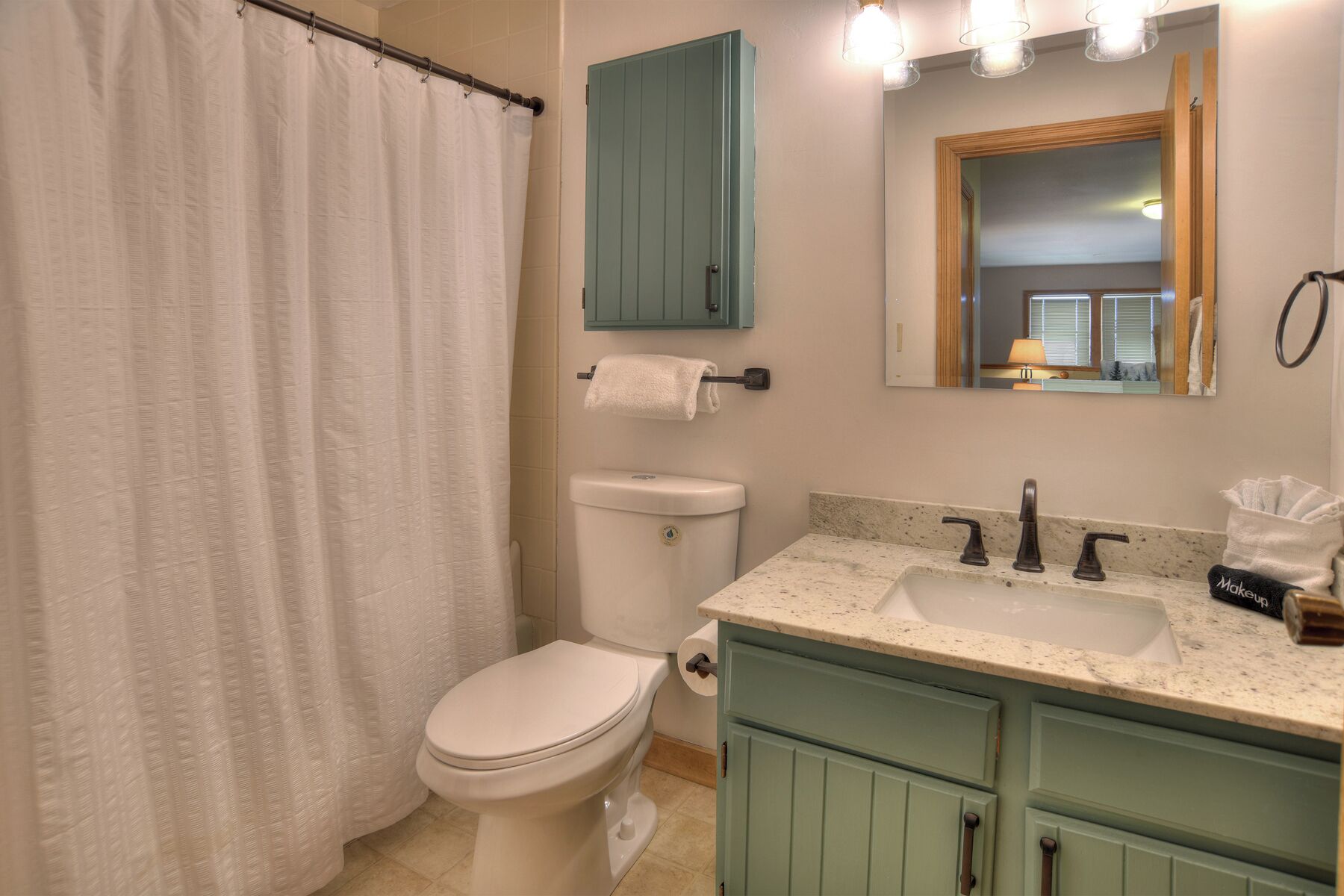 Un bagno con il water bianco, una tenda da doccia, un lavandino con uno specchio sopra e un asciugamano appeso al muro.