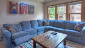 A living room with a blue sectional sofa, a wooden coffee table, and three abstract bear paintings on the wall. - The Blue Bear | Trails, Golf, Pool & Hot Tub - Near Town (Durango)