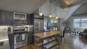 A modern kitchen with stainless steel appliances, a wooden dining table, and a loft-style living area with large windows.