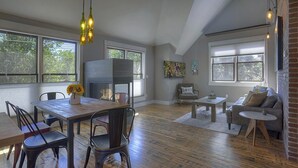 A spacious living room with a wooden floor, a dining area with a table and chairs, a fireplace, and large windows offering views of the outdoors.