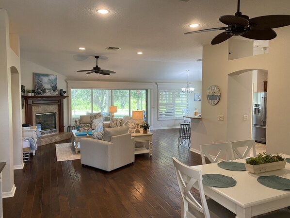 Uma sala de estar ampla com lareira, uma área de jantar com mesa e cadeiras, e uma cozinha com geladeira.