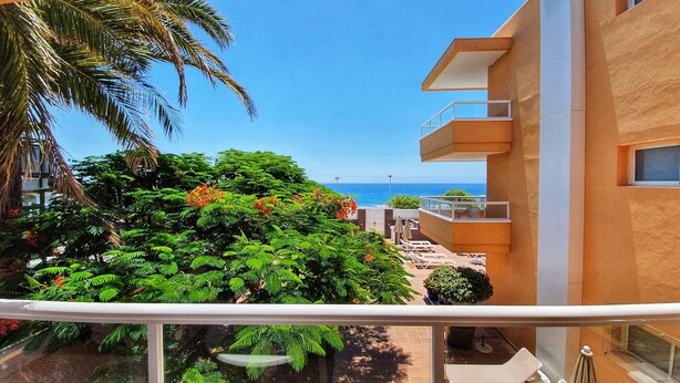 A balcony with a view of the ocean, lush greenery, and a modern building facade.