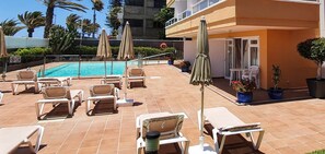A rooftop pool area with sun loungers and umbrellas, surrounded by potted plants and a building with a balcony.